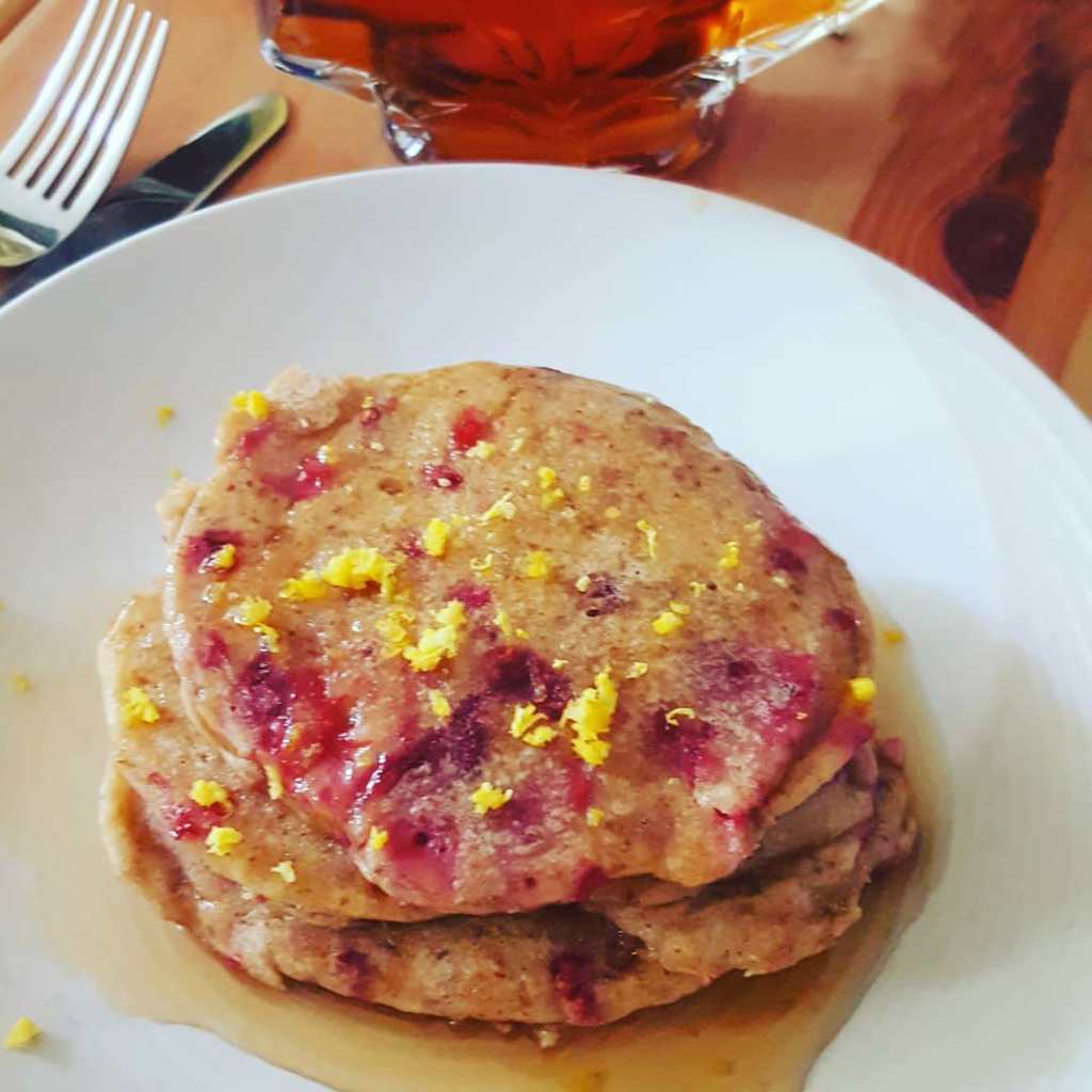 image: a stack of 3 raspberry lemon pancakes, soaking up maple syrup and sprinkled with fresh lemon zest on a white plate, rest on a wood coffee table next to a leaf-shaped bottle of maple syrup and a fork and knife.